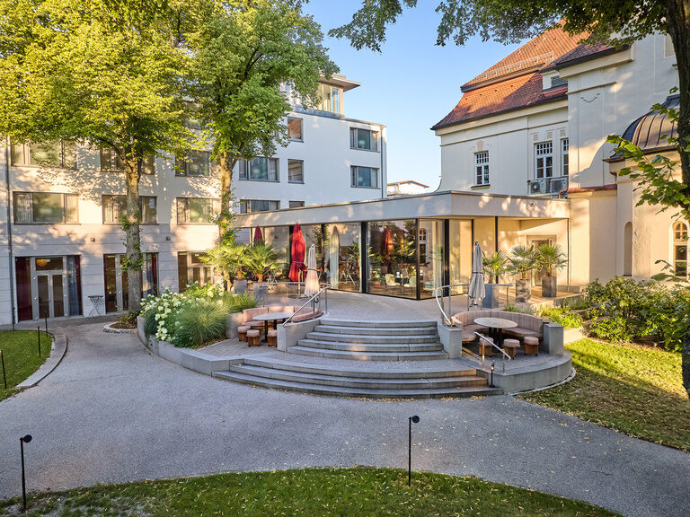 Blick von außen auf die Glasfront des Restaurant ASAM in Straubing, umgeben von üppiger grüner Bepflanzung. Im Hintergrund der Ostflügel des Hotels ASAM.
