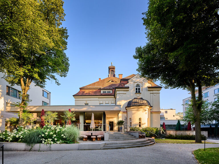Blick von außen auf die Glasfront des Restaurant ASAM in Straubing, umgeben von üppiger grüner Bepflanzung.