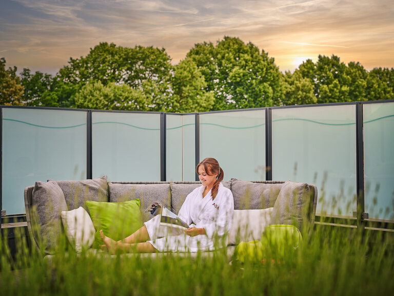 Dachterrasse des 4-Sterne-S Hotel Asam Straubing bei Abendstimmung mit Frau im Bademantel auf Outdoor-Couch beim Lesen. Dachterrasse des 4-Sterne-S Hotel Asam Straubing bei Abendstimmung mit Frau im Bademantel auf Outdoor-Couch beim Lesen.