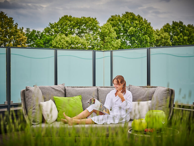 Entspannt auf der Dachterrasse: Frau im Bademantel liest auf der Outdoor-Couch, Bäume im Hintergrund, Hotel Asam Straubing.