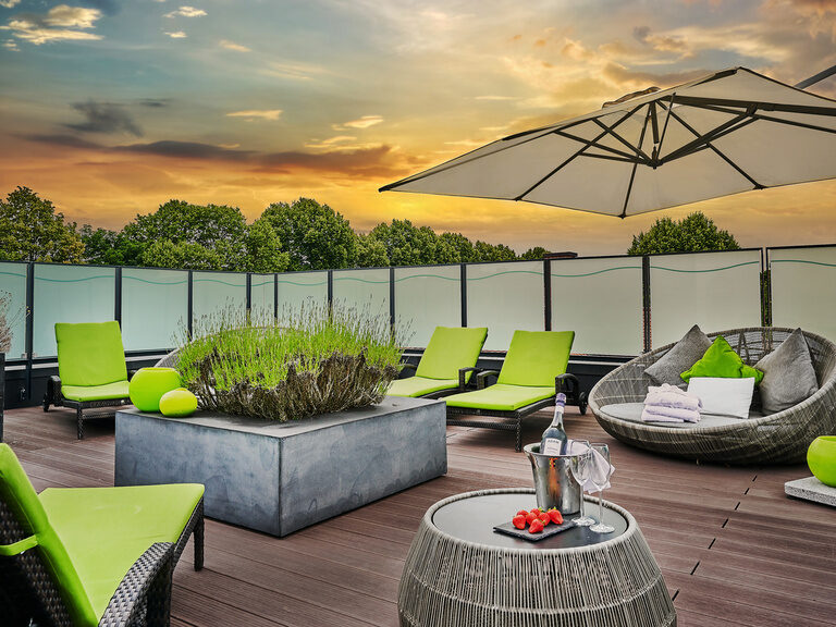 Dachterrasse im Wellnessbereich des Hotel Asam Straubing bei Abendstimmung mit Liegen, Sonnenschirm und Bäumen im Hintergrund.