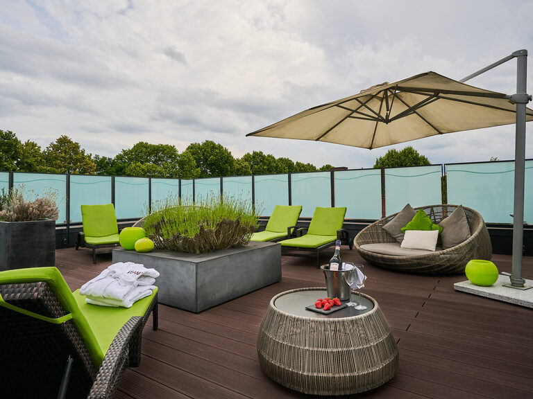 Begrünte Dachterrasse im Wellnessbereich des 4-Sterne-S Hotel Asam Straubing mit Liegen, Sonnenschirm und Bäumen im Sommer.