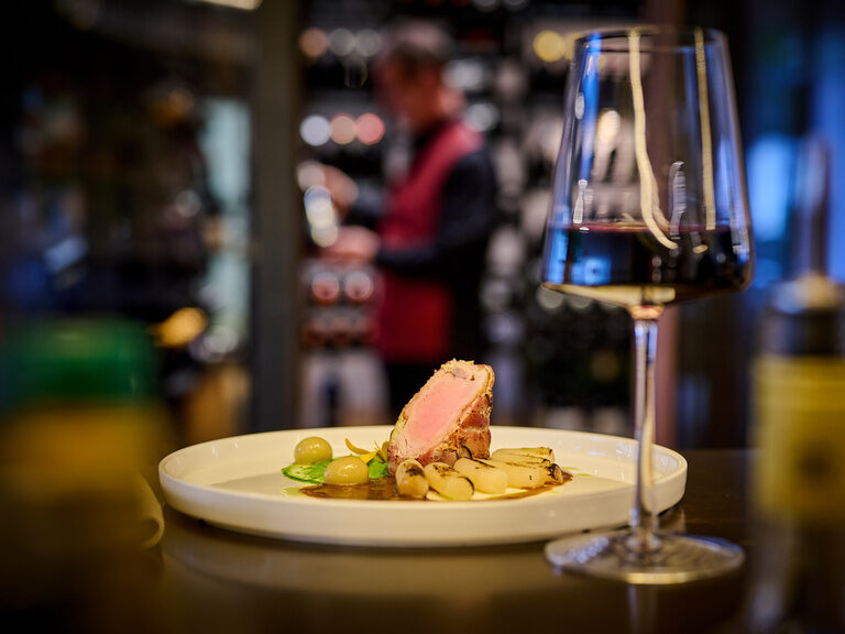 Ein fein angerichteter Telle rund ein Glas Rotwein im Restaurant ASAM, im Hintergrund der begehbare Weinschrank.