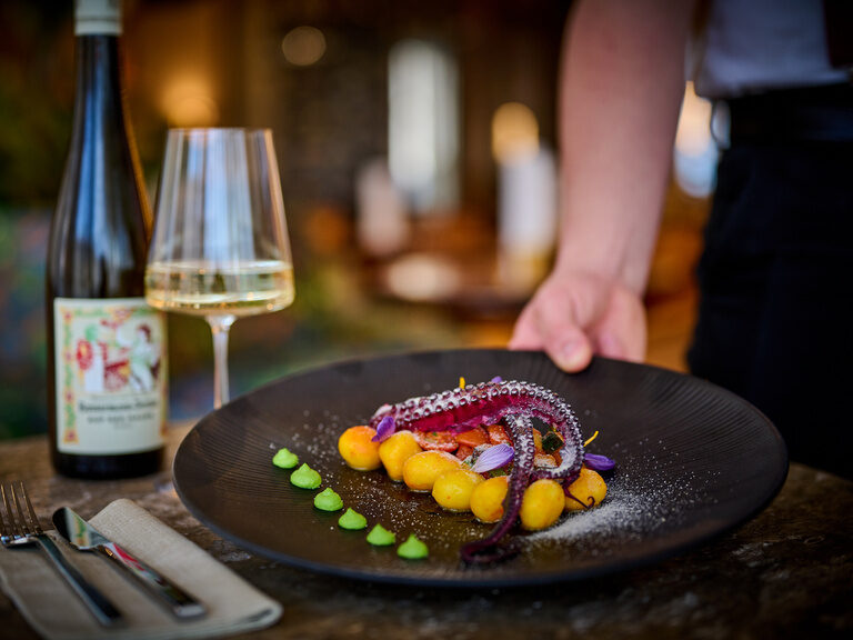 Ein Teller mit Oktopus wird gerade von einem Kellner serviert, ein Glas Wein steht bereits auf dem Tisch.