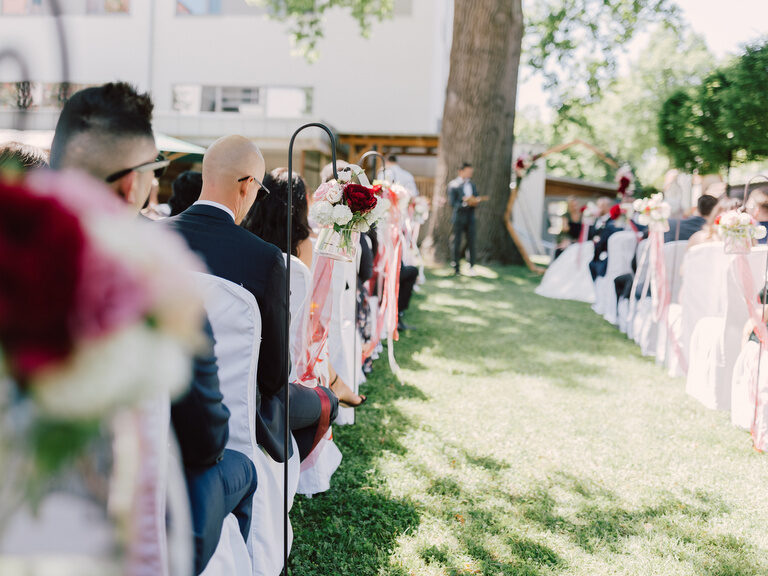 Blick auf eine Hochzeitsgesellschaft auf Stühlen mit weißen Hussen im Garten bei einer freien Trauung im Hotel ASAM, Straubing.