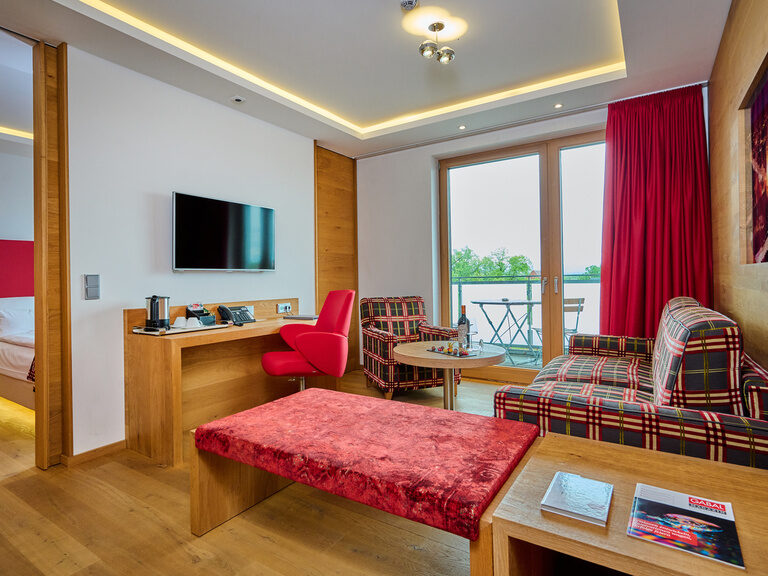 Wohnbereich der Suite mit Schlafcouch, Balkonzugang, Schreibtisch und abgetrenntem Schlafbereich, viel Holz und warme Farben. Wohnbereich der Suite mit Schlafcouch, Balkonzugang, Schreibtisch und abgetrenntem Schlafbereich, viel Holz und warme Farben.